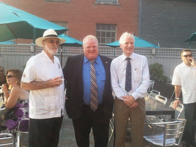 three men standing, two wearing white buttoned up shirts, the one in the middle wearing a blue dress shirt with a black blazer, grey fence in the back, teal umbrellas above the tables