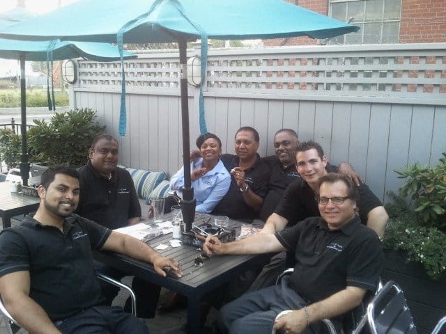 group of men wearing black gathering around black squared table, one women wearing a blue blouse, grey fence behind them, teal umbrella on top