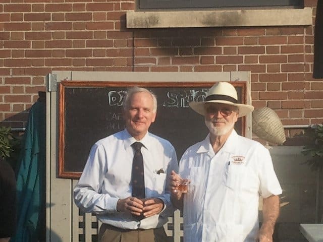 two men wearing white shirts, black chalkboard with brown border in the back, brown brick wall, outside
