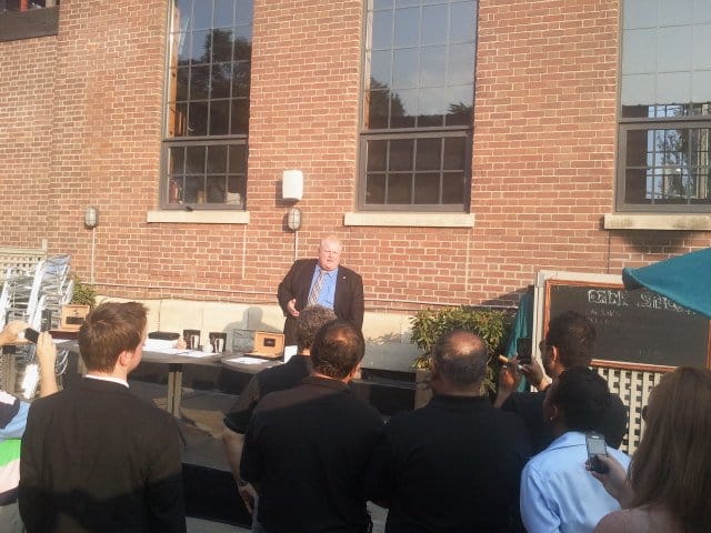 brown brick wall with three windows, man wearing blue dress shirt and striped tie with black jacket, group of 7 people standing in the front facing the wall
