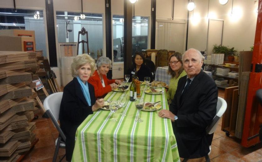 movie night, long green and white striped table cloth, five people nicely dressed sitting on white chairs, stacks of folded boxes on the side, white and red walls