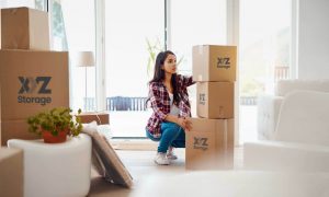 young woman wearing a plaid shirt with blue jeans and white shoes, packing her belongings using XYZ Storage boxes